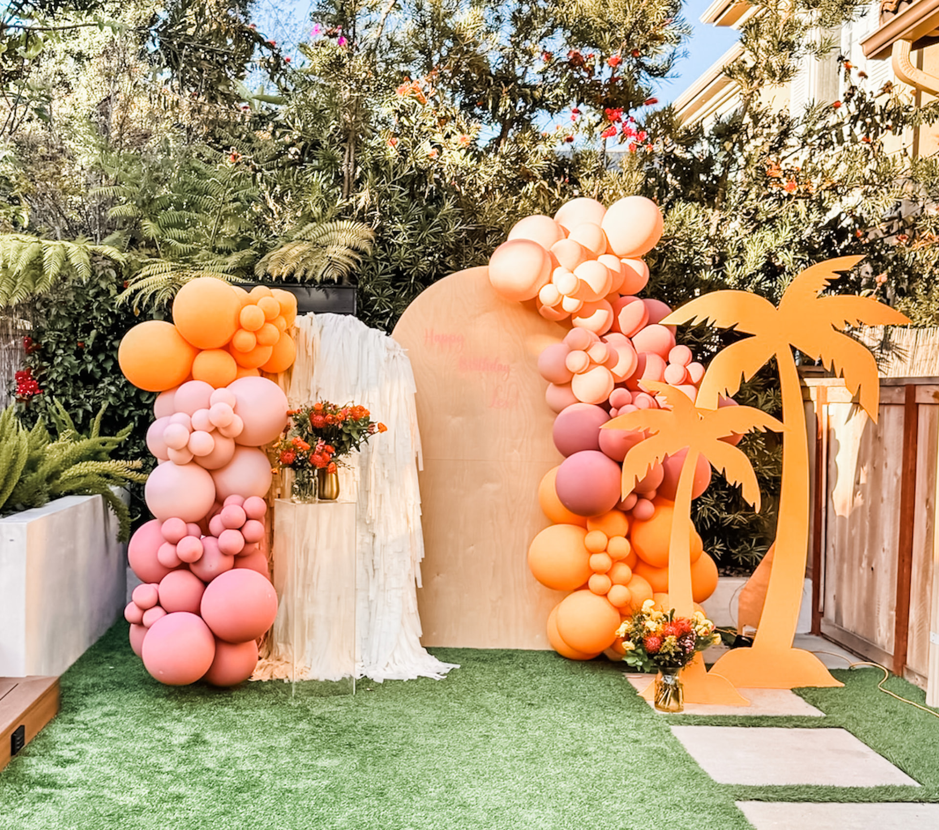 Peach, coral and apricot balloon garlands framing a wood arch backdrop with palm cutouts in a sunny garden